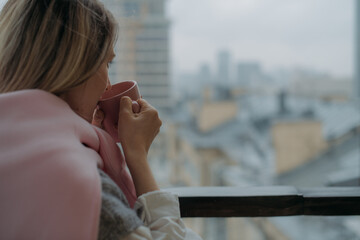 A young woman stands on the balcony of a city apartment with a mug of coffee.