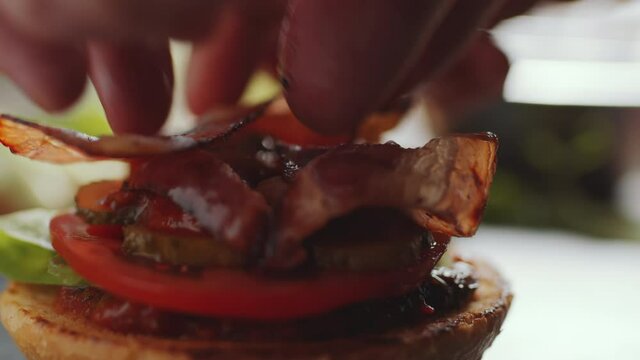 Close up shot of hands putting fried bacon slice on top of burger with lettuce, tomato and dill pickles