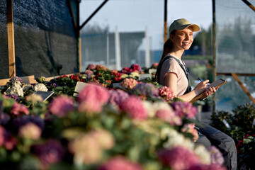 smiling positive female gardene writing in notebook in greenhouse, sit next to flowers.Good-looking Biotechnology woman engineer with clipboard and pen examining plant, outdoor portrait