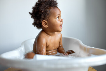 Small cute black child sitting at bath, washing, playing with soap and foam, indoors, copy space,...