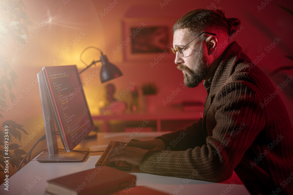 Poster side view of focused male programmer sitting at home during evening time and using modern pc for cod