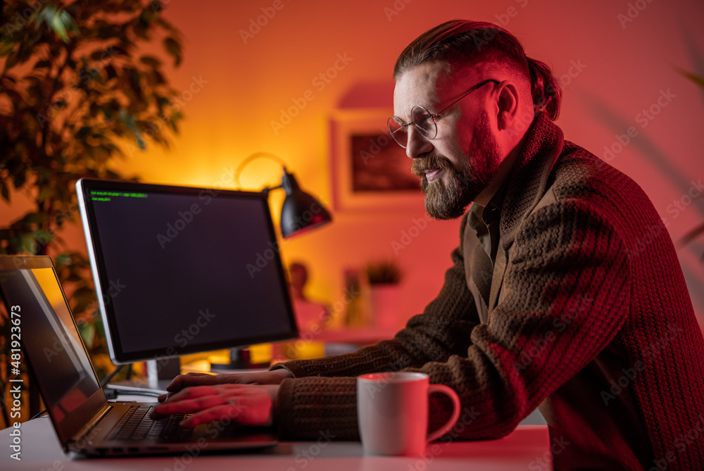 Poster positive bearded man in eyeglasses using modern computers for writing data code during night at home