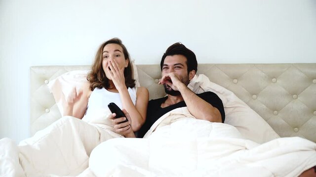 Lifestyle entertainment relationship concept. Cheerful young couple watching movie television while sitting together under blanket. Husband holding remote control tv in hand with own wife at home.