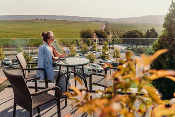 A middle-aged woman sits in a street cafe overlooking the mountains at sunset. She is dressed in a blue jacket and drinks coffee admiring the nature. Travel and vacation concept.