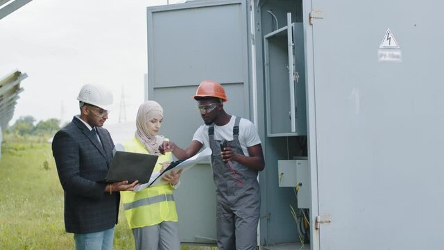 Indian man in suit and muslim woman in hijab using laptop and clipboard during examination of solar station. American technician checking serviceability of cables in switchgear. Power transformer.