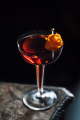 red boulevardier cocktail in hollow stem glass with orange peel rose garnish against black