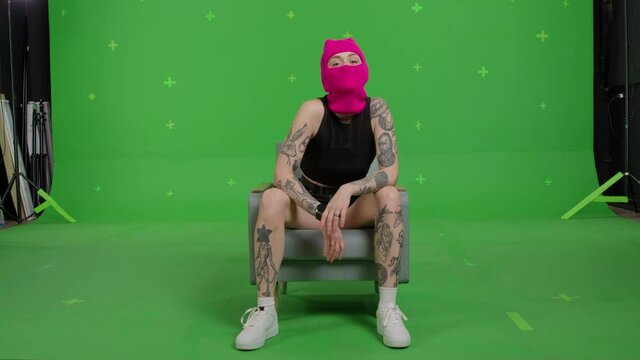 Young woman in pink balaclava sitting. Hooligan girl in mask looking at camera on green screen background . Chroma key . 4k uhd video