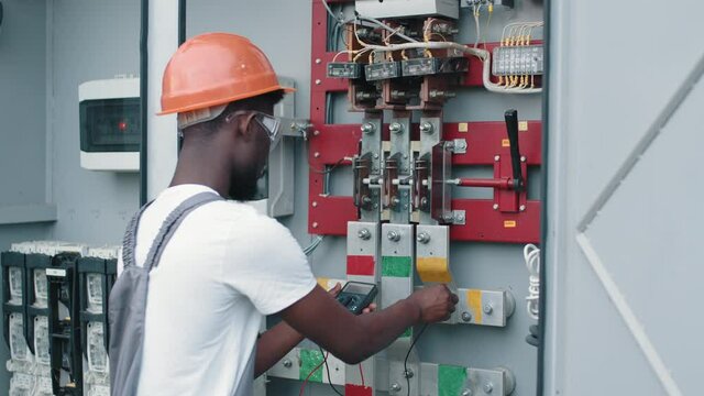 African american electrician in safety helmet and glasses checking voltage in switchgear while working at solar station. Repairing of modern equipment. Electrician checking voltage at solar station.