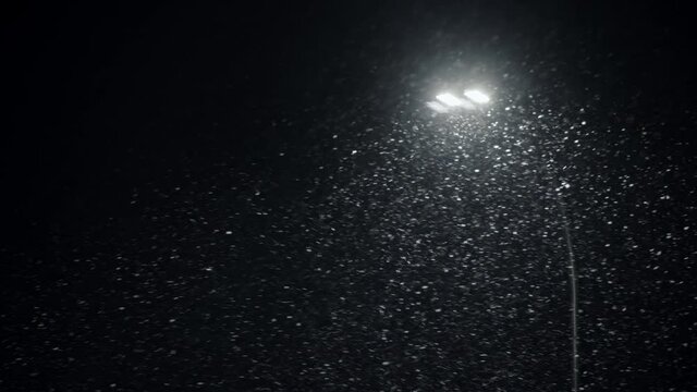Falling snow backlit with modern street lamp in city street at night in winter. Snowflakes illuminated by bright light in park. Snowfall, snow storm and and strong gusty wind