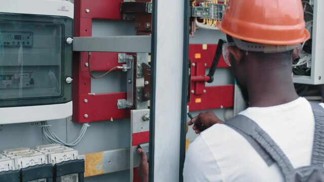 Close up of man using multimeter for work on solar station. African american electrician in safety helmet and glasses checking voltage in switchgear while working at solar station