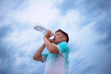 A boy against the sky drinks water from a plastic bottle. Thirst. Heat. Cloudy sky.