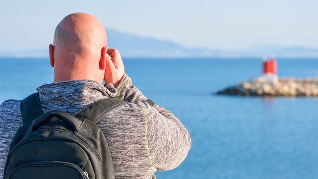 Rear view of a man with shaved head hair, taking photos of a seascape in the port