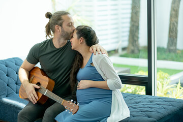 Fototapeta premium Man kiss on forehead of pregnant wife also hold guitar in living room after relax activity together and stay at home with love and happiness family.