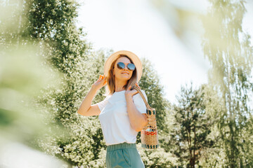 Pretty smiling caucasian young woman with freckles in straw hat, glasses and handbag looking away while standing in nature on sunny summer day. Portrait of happy charming girl walking outdoors