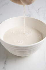 Nut milk bag squeezing milk into a white bowl with almond milk placed on a white marble surface - obrazy, fototapety, plakaty
