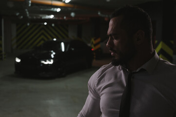 strong rich young bearded man in white shirt with black tie in underground garage with luxury sport...