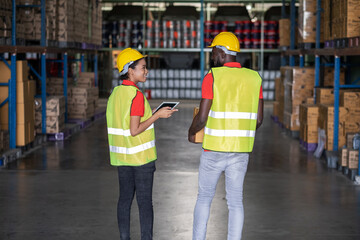 Back view of Africa American engineer woman holding tablet wearing safety helmet and vest standing in the automotive part warehouse. Looking at friend. Portrait of worker. Logistic and business.