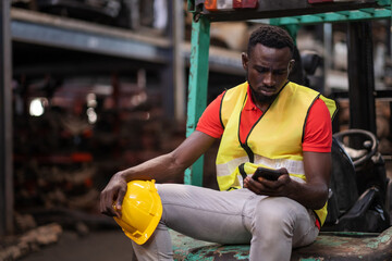 Africa American holding safety helmet and sitting at forklift in the automotive part warehouse. Worker look at smartphone and get the bad news. Feeling stress from fired from work. Disappoint