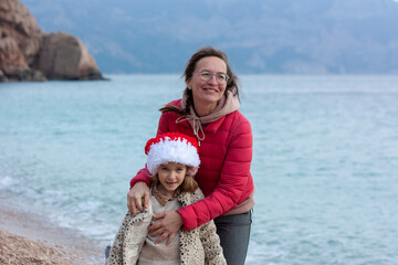 Mom and daughter in Santa's hat are walking along the seashore, they are laughing and talking