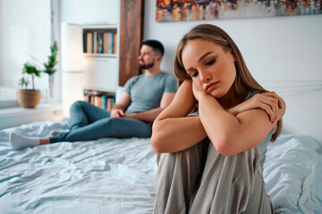 Upset young woman in home clothes sitting on the bed, in the background sitting man. Family troubles. - obrazy, fototapety, plakaty