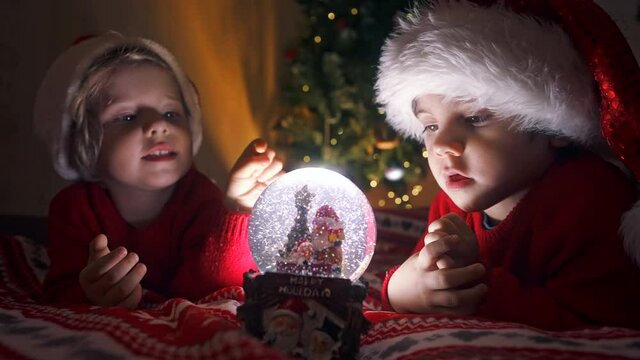 Cute little boys in Santa hats with snow globe. New Year's gift toy for children. Magic Christmas time, toddlers look at glass ball with interest. Family, brothers, holidays and celebration concept
