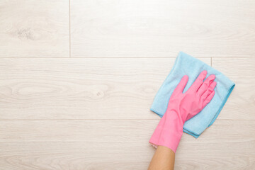Young adult woman hand in pink rubber protective gloves using blue dry rag and wiping light laminate floor surface in room at home. Closeup. Empty place for text. Top down view.