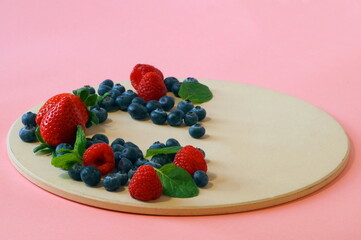 Fresh berries - strawberries, raspberries and blueberries and fragrant mint leaves - are neatly stacked on a round tray in a crescent shape. Photography background - pink