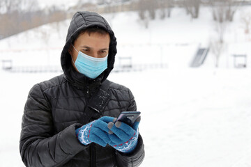 Fototapeta na wymiar Coronavirus protection, man in medical mask on a winter city street with smartphone in hands. Concept of safety during covid-19 pandemic, snow weather
