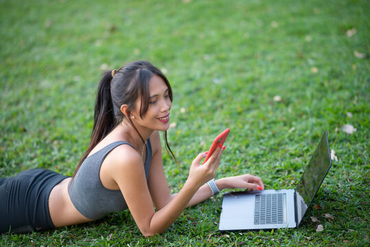 Young and beautiful asian woman lying on the park grass using laptop and smartphone.