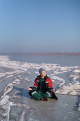 Fototapeta na wymiar A woman in a colorful thermal costume enjoying winter while sitting in a meditation pose on a frozen lake with blue sky in the background. Freedom, peace, quiet, serenity