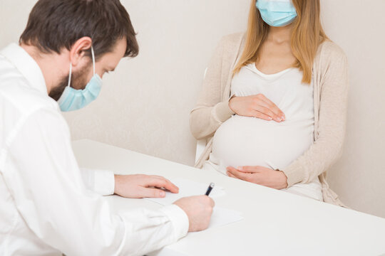 Doctor man sitting at table and writing records about young adult pregnant woman. Regular examination in pregnancy time. People using face protective masks at gynecological office.