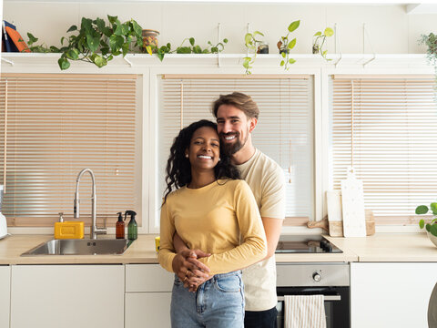 Multiethnic heterosexual couple smiling hugging and looking at camera at home