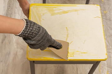 a hand with a paint scraper removes a layer of paint from a stool