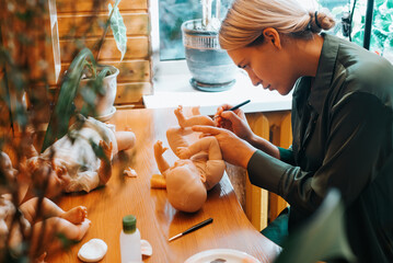 Young female puppeteer decorating reborn sitting at table in home workshop. Side view of girl...