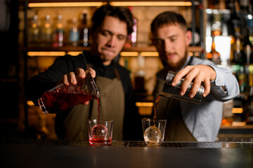 two glasses with ice in which bartenders carefully pour different cocktails