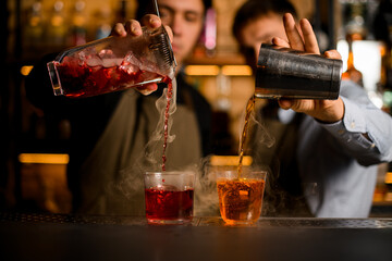view of two transparent glasses with ice, into which bartenders carefully pour various steaming cocktails
