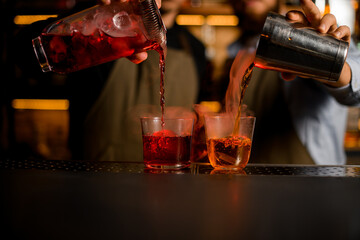 two transparent glasses with ice, into which bartenders carefully pour various bright steaming cocktails