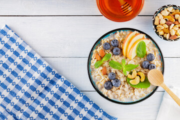 Oatmeal bowl and honey on a white boards. Oatmeal porridge with blueberries, apple slices and fresh mint. Healthy breakfast. Top view. Copy space