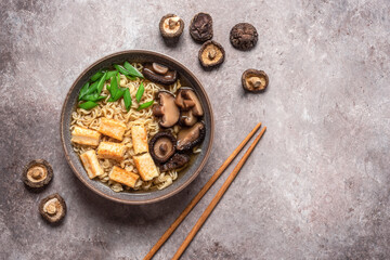 Asian vegan ramen noodle soup with roasted tofu cheese and shiitake mushrooms in a bowl on brown grunge background. Top view. Selective Focus.