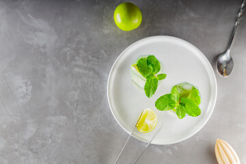 Mojito cocktail and bar accessories on gray background. Two highball glasses of mojito on a white plate. Summer refreshing cocktail with lime and mint. Copy space