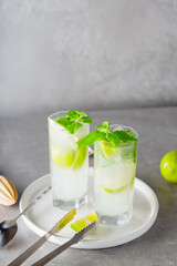 Mojito cocktail and bar accessories on gray background. Two highball glasses of mojito on a white plate. Summer refreshing cocktail with lime and mint. Copy space