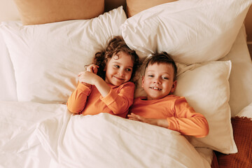 Obraz na płótnie Canvas Smiling funny twin children a little boy and a girl in pajamas sleep together lying on pillows on the bed in a cozy comfortable bedroom. Top view. Selective focus