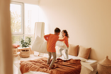 Happy cheerful sleepy children brother and sister in orange bright pajamas have fun laughing and fighting with pillows in a cozy bedroom at home. Children's pajama party. Selective focus