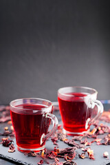 Red hibiscus tea in glass mug on a dark background. Cup of hibiscus tea and dry hibiscus petals on a slate board. Copy space