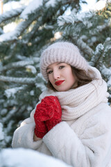 Portrait of beautiful girl in red mittens and white hat on snow-covered fir trees background. Vertical frame. Copy space