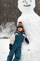 Portrait of boy in knitted hat and overalls next to large snowman. Child playing with snow man.