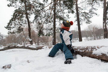 Fototapeta na wymiar Little boy sitting on log in winter forest. Child in knitted hat and winter jumpsuit looks up at sky