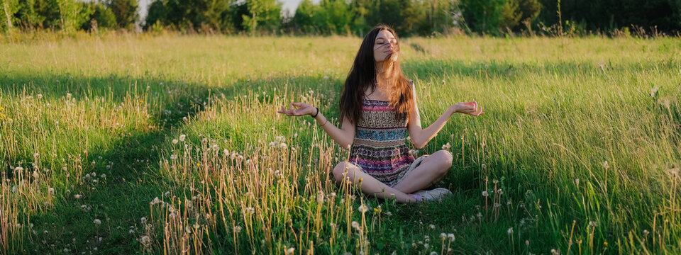 Young woman practicing yoga sitting in easy pose outdoors in green field. Unity with nature concept.