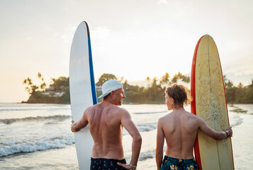 Father with teenager son standing with surfboards on the sandy ocean beach with palm trees on background lightened with sunset sun. They smiling and have a conversation. Family active vacation concept