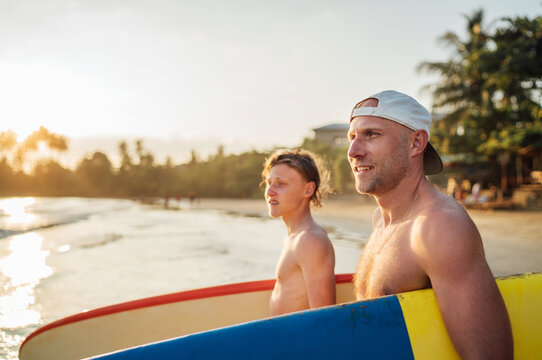 Smiling man with Young teen boy son with surfboards go to the sea for surfing. They have a winter vacation and enjoying a beautiful sunset light. Family active vacation concept.